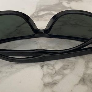 Puede incluir: Gafas de sol negras con lentes oscuras. Las gafas tienen una montura negra gruesa y brillante y est&aacute;n sobre una superficie de m&aacute;rmol. Las patillas de las gafas est&aacute;n plegadas. El nombre de la marca es visible en la patilla.