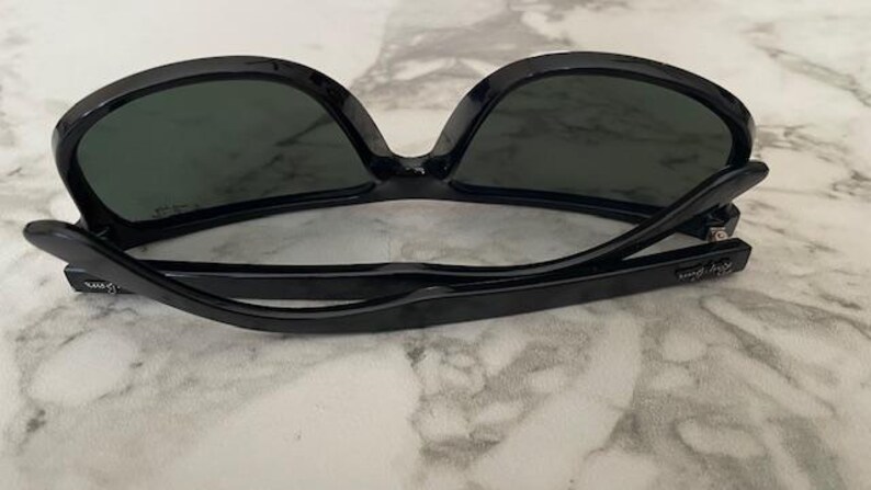 Puede incluir: Gafas de sol negras con lentes oscuras. Las gafas tienen una montura negra gruesa y brillante y est&aacute;n sobre una superficie de m&aacute;rmol. Las patillas de las gafas est&aacute;n plegadas. El nombre de la marca es visible en la patilla.