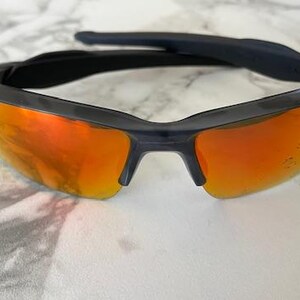 Peut inclure: Lunettes de soleil noires avec des verres orange. Les lunettes ont un design moderne avec une monture noire et une barre sup&eacute;rieure incurv&eacute;e. Les verres sont d'une couleur orange vif, refl&eacute;tant la lumi&egrave;re.