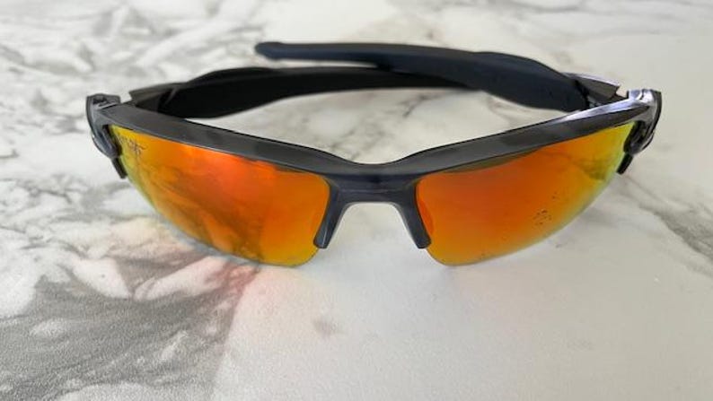 Peut inclure: Lunettes de soleil noires avec des verres orange. Les lunettes ont un design moderne avec une monture noire et une barre sup&eacute;rieure incurv&eacute;e. Les verres sont d'une couleur orange vif, refl&eacute;tant la lumi&egrave;re.