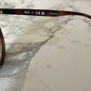 May include: Tortoiseshell sunglasses with brown lenses. The frame has a classic design with a gradient color pattern. The Ray-Ban logo is visible on the temple arm. Made in China is printed on the arm.