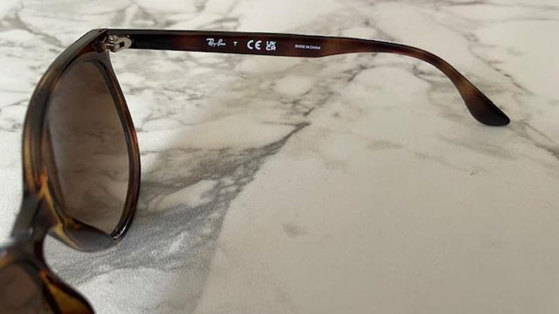 May include: Tortoiseshell sunglasses with brown lenses. The frame has a classic design with a gradient color pattern. The Ray-Ban logo is visible on the temple arm. Made in China is printed on the arm.
