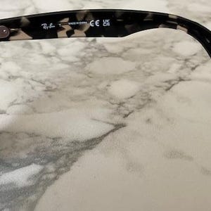 Puede incluir: Gafas de sol con montura moteada en blanco y negro y lentes azules. La patilla tiene el logotipo de Ray-Ban y el texto "MADE IN CHINA" y "CE UK". Las gafas de sol est&aacute;n sobre una superficie de m&aacute;rmol.