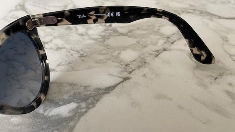 Puede incluir: Gafas de sol con montura moteada en blanco y negro y lentes azules. La patilla tiene el logotipo de Ray-Ban y el texto "MADE IN CHINA" y "CE UK". Las gafas de sol est&aacute;n sobre una superficie de m&aacute;rmol.