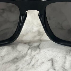May include: Close-up of black sunglasses with dark gray lenses. The sunglasses have a thick, rectangular frame and a bridge connecting the two lenses. The lenses are slightly reflective.