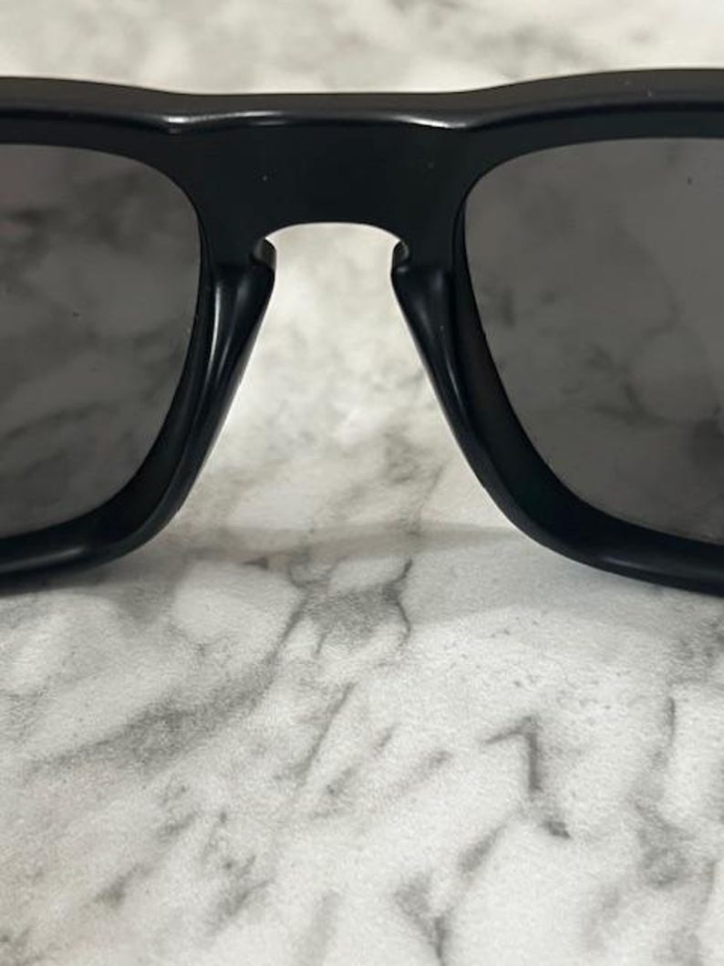 May include: Close-up of black sunglasses with dark gray lenses. The sunglasses have a thick, rectangular frame and a bridge connecting the two lenses. The lenses are slightly reflective.