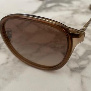 May include: Brown-tinted sunglasses with a gold-colored frame. The lenses have a gradient tint, transitioning from a darker shade at the top to a lighter shade at the bottom. The glasses are set against a marble-like surface.