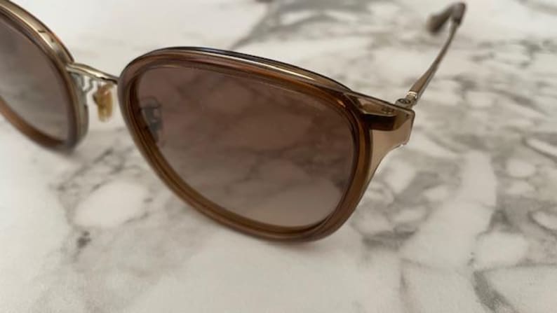 May include: Brown-tinted sunglasses with a gold-colored frame. The lenses have a gradient tint, transitioning from a darker shade at the top to a lighter shade at the bottom. The glasses are set against a marble-like surface.