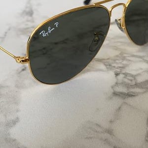 May include: Gold-rimmed aviator sunglasses with dark lenses. The Ray-Ban logo is visible on the lens. The sunglasses have a classic design with a double bridge and thin temples. The sunglasses are resting on a marble surface.
