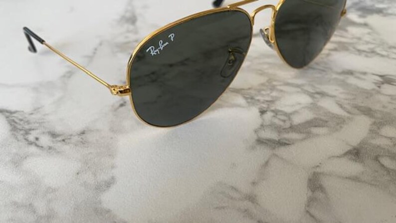 May include: Gold-rimmed aviator sunglasses with dark lenses. The Ray-Ban logo is visible on the lens. The sunglasses have a classic design with a double bridge and thin temples. The sunglasses are resting on a marble surface.