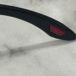 May include: Close-up of the temple of black sunglasses. The temple has a red rectangular accent with the word "USA" printed on it. The sunglasses are on a white surface.