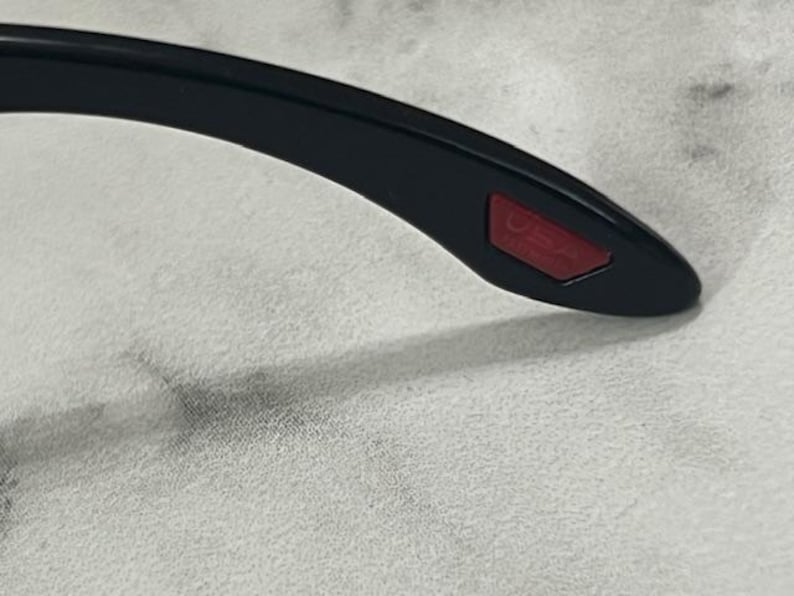 May include: Close-up of the temple of black sunglasses. The temple has a red rectangular accent with the word "USA" printed on it. The sunglasses are on a white surface.
