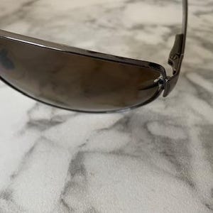 May include: A pair of sunglasses with a dark brown tint and a silver frame. The sunglasses have a sleek, modern design with a curved shape. The lenses are a gradient color, transitioning from dark to light.