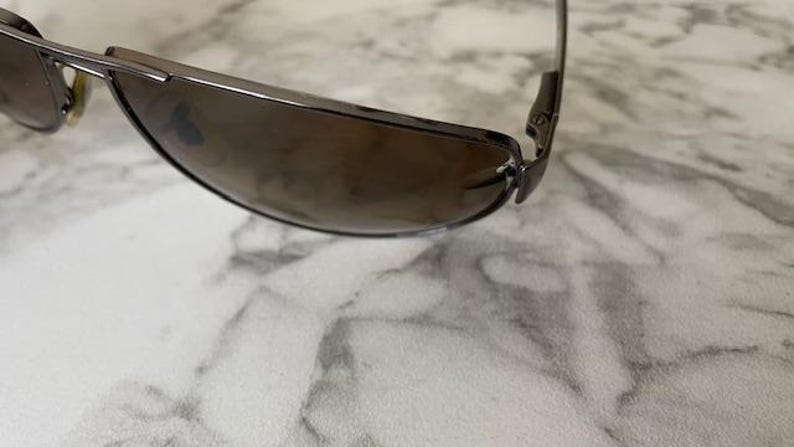 May include: A pair of sunglasses with a dark brown tint and a silver frame. The sunglasses have a sleek, modern design with a curved shape. The lenses are a gradient color, transitioning from dark to light.