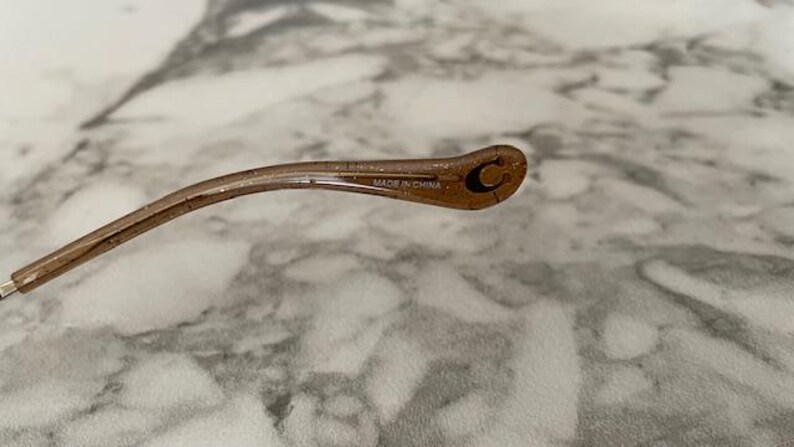 May include: A close-up of the arm of a pair of eyeglasses. The arm is a translucent brown color with glitter and a small "C" logo. The text "MADE IN CHINA" is printed on the arm.
