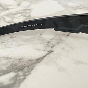 Puede incluir: Primer plano de gafas de sol negras con lentes azules. La patilla de la montura presenta el texto "OO9239-3060 60 18 132 3P". Las gafas de sol est&aacute;n sobre una superficie similar al m&aacute;rmol.