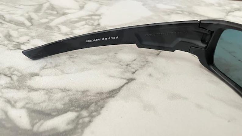 Puede incluir: Primer plano de gafas de sol negras con lentes azules. La patilla de la montura presenta el texto "OO9239-3060 60 18 132 3P". Las gafas de sol est&aacute;n sobre una superficie similar al m&aacute;rmol.