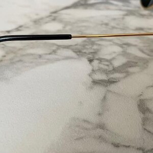 May include: Close-up of sunglasses with gold-colored frames and dark lenses. The temple arm is thin and gold, with a black earpiece. The sunglasses are set against a marble-patterned surface.