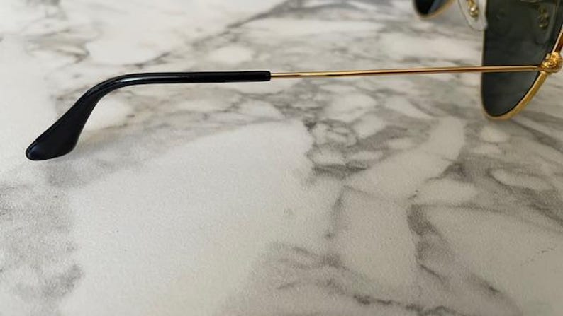 May include: Close-up of sunglasses with gold-colored frames and dark lenses. The temple arm is thin and gold, with a black earpiece. The sunglasses are set against a marble-patterned surface.