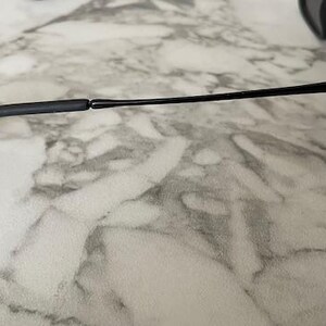 Puede incluir: Gafas de sol negras con un dise&ntilde;o elegante y moderno. Las monturas son de color negro mate y las lentes son oscuras. Las patillas son finas y se extienden hasta los terminales. Las gafas de sol est&aacute;n sobre una superficie de m&aacute;rmol.