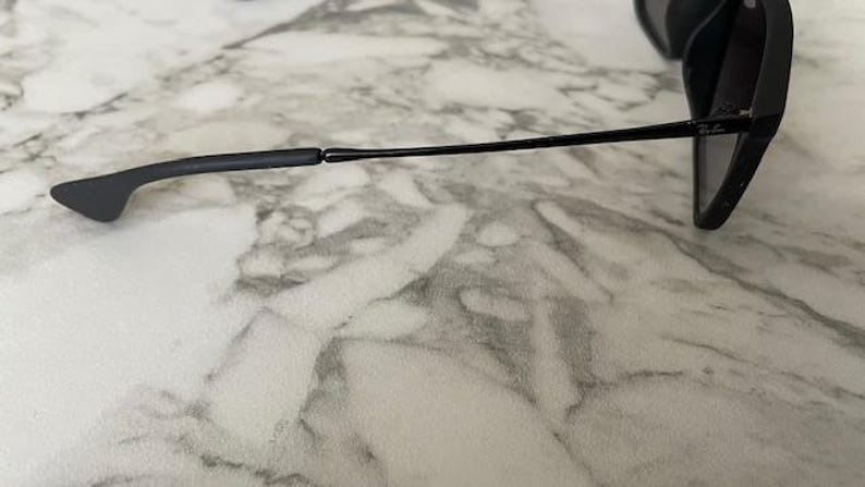 Puede incluir: Gafas de sol negras con un dise&ntilde;o elegante y moderno. Las monturas son de color negro mate y las lentes son oscuras. Las patillas son finas y se extienden hasta los terminales. Las gafas de sol est&aacute;n sobre una superficie de m&aacute;rmol.