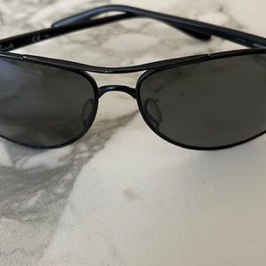 May include: Black sunglasses with a sleek, modern design. The sunglasses feature a black frame and dark lenses. The lenses are oval-shaped and the frame has a double bridge. The sunglasses are resting on a white and gray marble surface.
