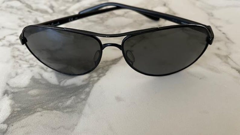 May include: Black sunglasses with a sleek, modern design. The sunglasses feature a black frame and dark lenses. The lenses are oval-shaped and the frame has a double bridge. The sunglasses are resting on a white and gray marble surface.