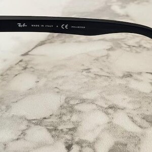 Puede incluir: Primer plano de gafas de sol negras con lente verde oscuro. La patilla tiene el logotipo de Ray-Ban, "MADE IN ITALY", "CE" y "POLARIZED" impresos. Las gafas de sol est&aacute;n sobre una superficie de m&aacute;rmol.