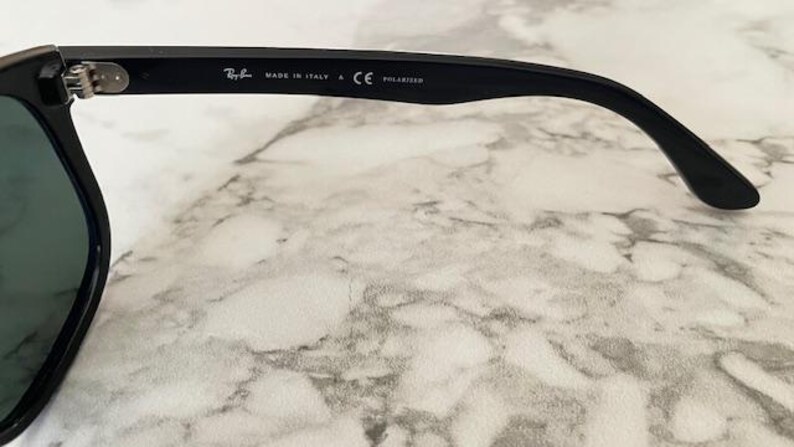 Puede incluir: Primer plano de gafas de sol negras con lente verde oscuro. La patilla tiene el logotipo de Ray-Ban, "MADE IN ITALY", "CE" y "POLARIZED" impresos. Las gafas de sol est&aacute;n sobre una superficie de m&aacute;rmol.