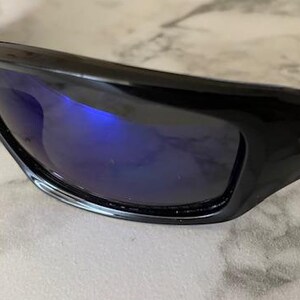 May include: Close-up of black sunglasses with blue reflective lenses. The sunglasses have a glossy finish and a curved design. The lenses reflect the surrounding environment, creating a vibrant blue hue.