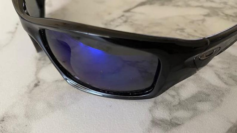 May include: Close-up of black sunglasses with blue reflective lenses. The sunglasses have a glossy finish and a curved design. The lenses reflect the surrounding environment, creating a vibrant blue hue.