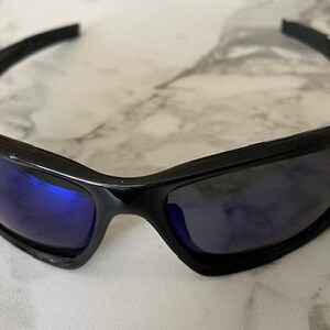May include: Black sunglasses with a glossy finish and blue-tinted lenses. The sporty eyewear features a curved frame and a streamlined design, suitable for outdoor activities. The sunglasses are designed to provide eye protection from the sun.