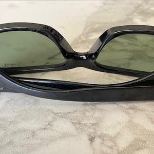 Puede incluir: Gafas de sol negras con lentes verdes, apoyadas sobre una superficie clara. Las gafas tienen una montura rectangular cl&aacute;sica con el logotipo de Ray-Ban visible en la patilla. Las lentes son de un verde apagado.