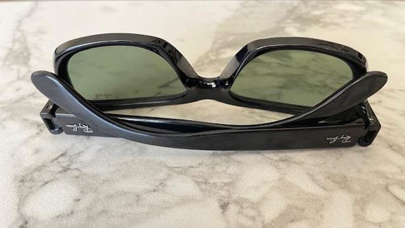 Puede incluir: Gafas de sol negras con lentes verdes, apoyadas sobre una superficie clara. Las gafas tienen una montura rectangular cl&aacute;sica con el logotipo de Ray-Ban visible en la patilla. Las lentes son de un verde apagado.