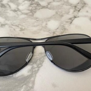 May include: Black aviator sunglasses with dark gray lenses. The sunglasses have a black metal frame and a subtle blue accent on the temples. The lenses are a dark gray color.
