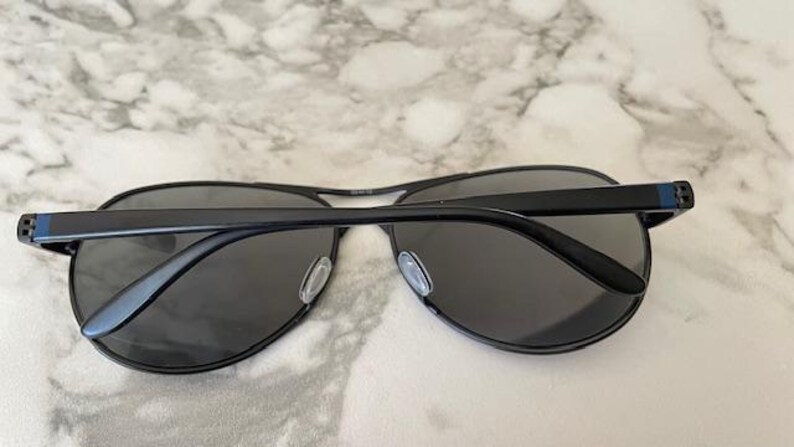 May include: Black aviator sunglasses with dark gray lenses. The sunglasses have a black metal frame and a subtle blue accent on the temples. The lenses are a dark gray color.