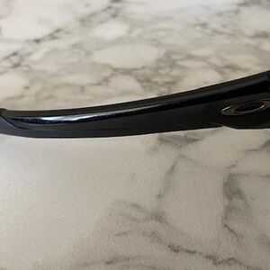 May include: Close-up of black sunglasses with a curved frame and a dark gray logo on the temple. The sunglasses have a sleek, modern design, and the lenses are not visible. The frame is made of a glossy material.
