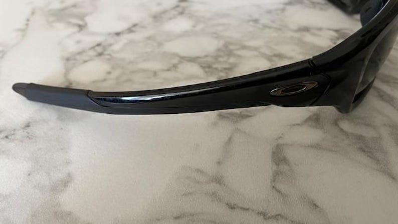 May include: Close-up of black sunglasses with a curved frame and a dark gray logo on the temple. The sunglasses have a sleek, modern design, and the lenses are not visible. The frame is made of a glossy material.