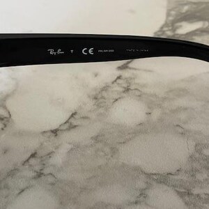 Puede incluir: Primer plano de la patilla de unas gafas de sol negras. El logotipo de Ray-Ban y otro texto son visibles en la patilla. Las gafas de sol est&aacute;n sobre una superficie de m&aacute;rmol blanco y gris. Las gafas de sol est&aacute;n polarizadas y fabricadas en EE. UU.