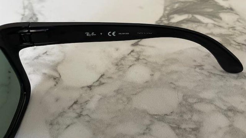 Puede incluir: Primer plano de la patilla de unas gafas de sol negras. El logotipo de Ray-Ban y otro texto son visibles en la patilla. Las gafas de sol est&aacute;n sobre una superficie de m&aacute;rmol blanco y gris. Las gafas de sol est&aacute;n polarizadas y fabricadas en EE. UU.