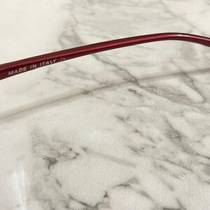 May include: Close-up of a pair of red sunglasses. The frame is a deep red color, with the text "MADE IN ITALY" printed on the temple. The lenses are a dark gray color.
