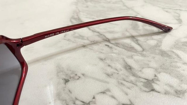 May include: Close-up of a pair of red sunglasses. The frame is a deep red color, with the text "MADE IN ITALY" printed on the temple. The lenses are a dark gray color.