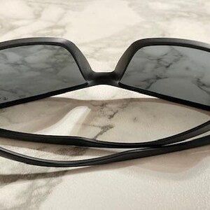 May include: Black sunglasses with gray lenses. The sunglasses have a matte black frame and a sleek, modern design. The arms are thin and curve down, with silver accents near the hinges. The lenses are rectangular.