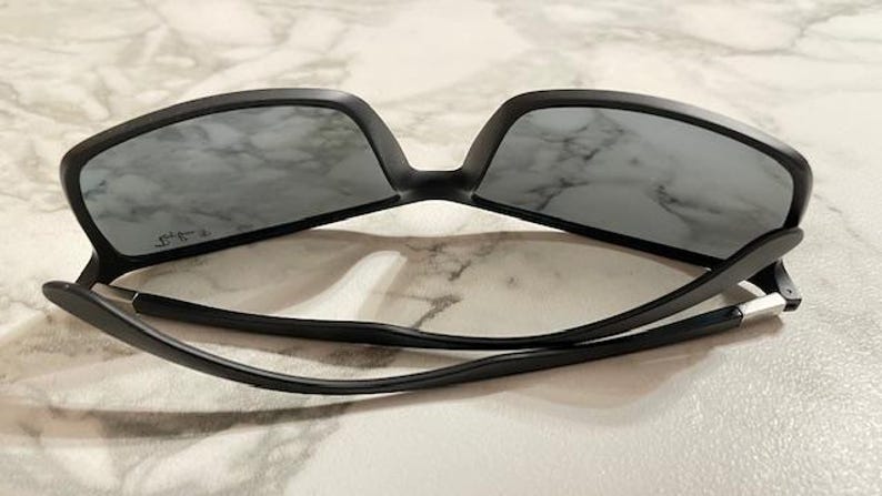 May include: Black sunglasses with gray lenses. The sunglasses have a matte black frame and a sleek, modern design. The arms are thin and curve down, with silver accents near the hinges. The lenses are rectangular.
