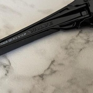 May include: Close-up of black sunglasses with the word "VALVE" printed on the arm. The arm also has the numbers "OO9236-1260 80 16 133 3P" printed on it. The sunglasses are on a marble surface.