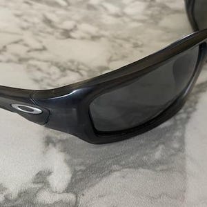 May include: Black sunglasses with dark lenses. The sunglasses have a thick, curved frame and a silver logo on the temple. The glasses are resting on a white and gray marble surface.