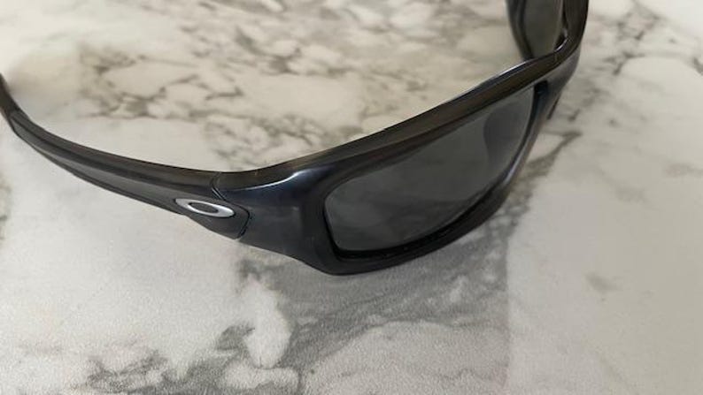 May include: Black sunglasses with dark lenses. The sunglasses have a thick, curved frame and a silver logo on the temple. The glasses are resting on a white and gray marble surface.