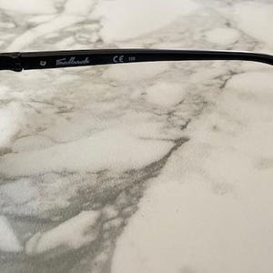 May include: Black sunglasses with round lenses. The frame is black with the text "Fallback" and "CE 138" printed on the temple. The sunglasses are set against a marble background.