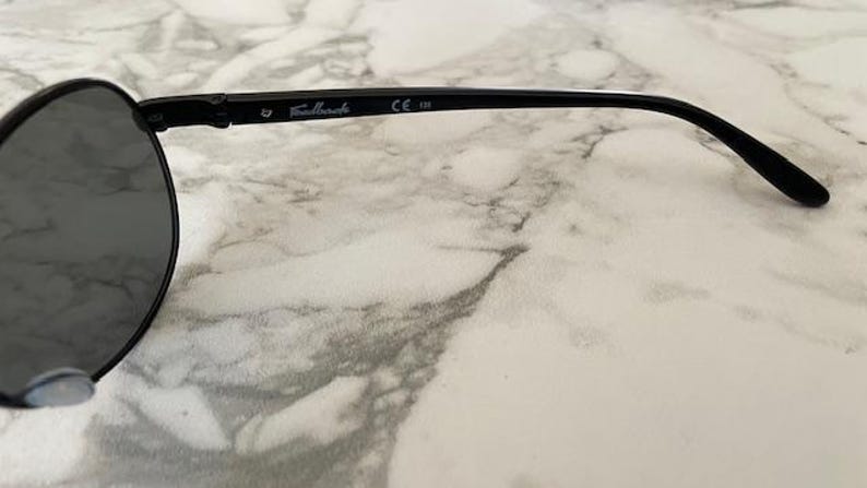 May include: Black sunglasses with round lenses. The frame is black with the text "Fallback" and "CE 138" printed on the temple. The sunglasses are set against a marble background.