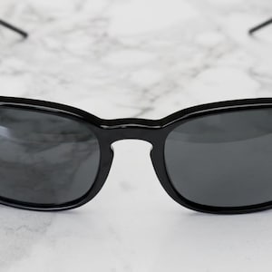 Puede incluir: Gafas de sol negras con lentes grises oscuras. Las gafas tienen una montura redondeada y patillas finas. El logotipo de Ray-Ban es visible en la lente izquierda. Las gafas est&aacute;n sobre un fondo de m&aacute;rmol blanco y gris.